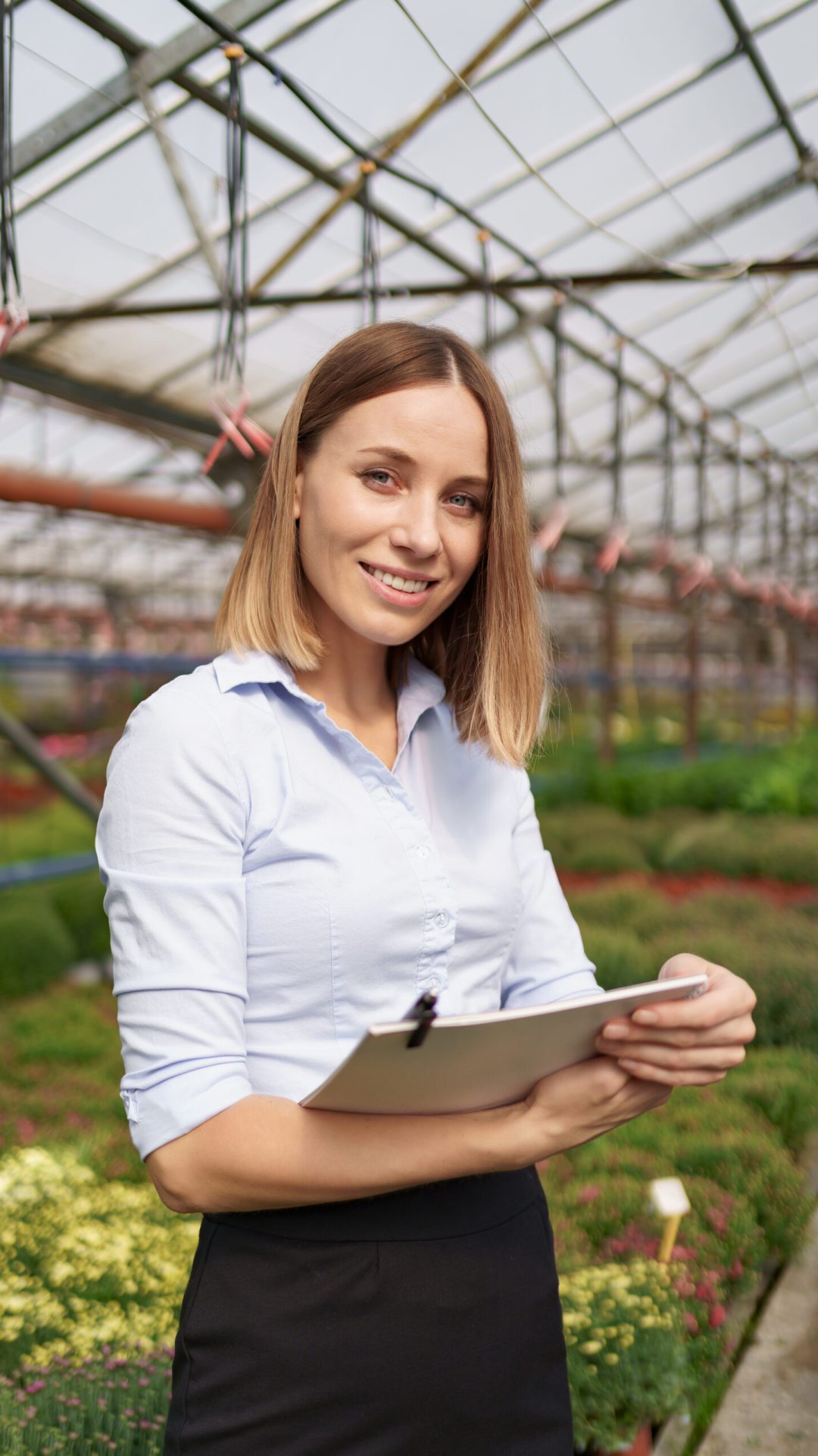 Smiling Greenhouse owner posing with a notepad in her hands having many flowers in background and glass roof